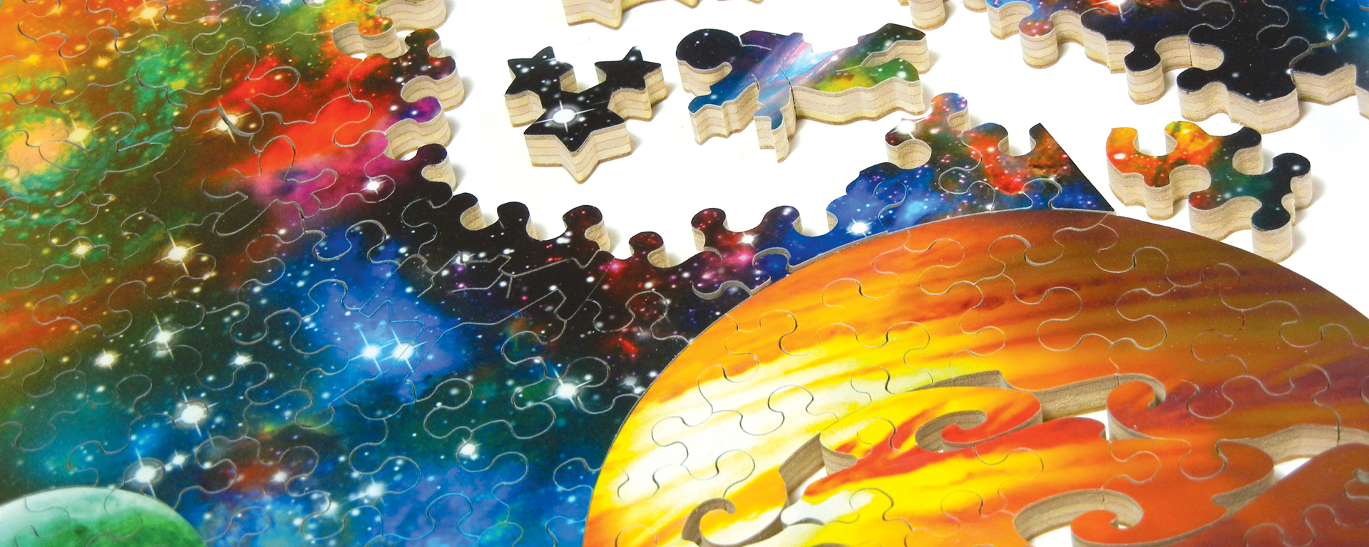Wooden space puzzle in progress featuring Jupiter in the foreground and stars, galaxies, and nebulae with whimsy shaped pieces such as a cluster of stars and an astronaut.